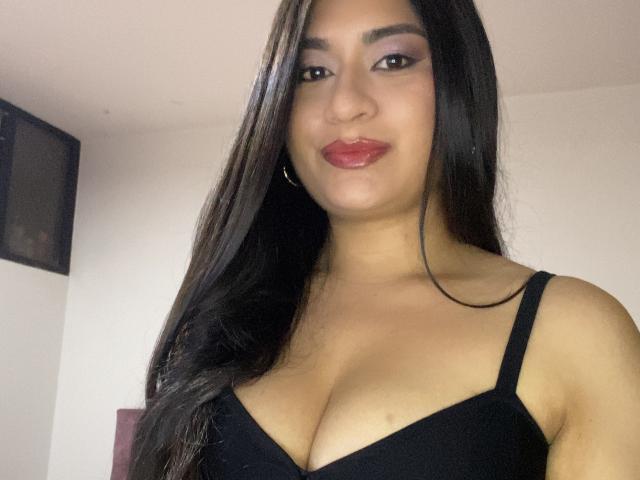 MiaDuboisX with long black hair, tan skin, and red lipstick, wearing a black bra that shows her large, shiny tits. She's smiling with hoop earrings and a subtle nose.