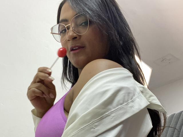 CameronJones with long black hair, glasses, and a pink bra, holding a red lollipop, wearing a white shirt off her shoulder, looking at the camera. Boobs visible.