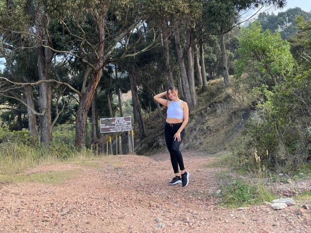 EliBaker, with a medium butt, wearing a white sports bra and black leggings, stands on a gravel path, touching her head, with trees and greenery in the background.