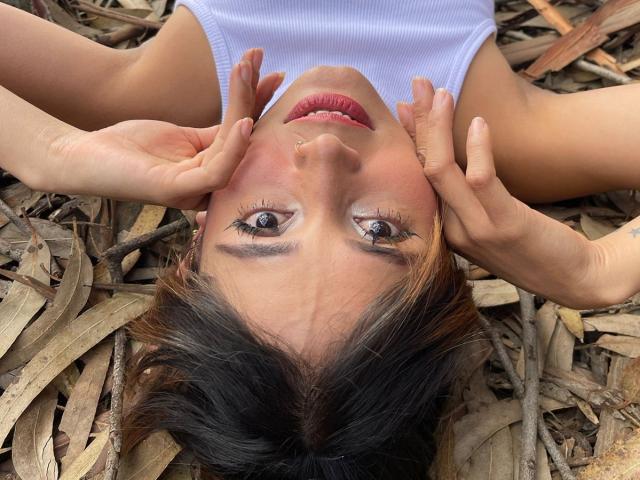 EliBaker's face is upside down with hands on cheeks, brown eyes, red lipstick, white tank top, and brown wavy hair on wooden leaves. EliBaker's nose is pierced.