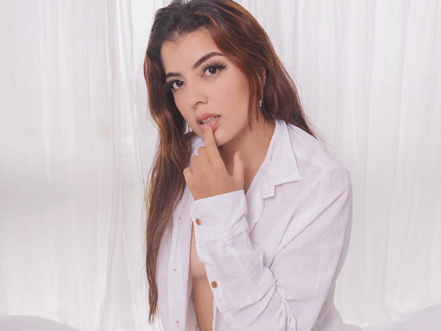 KymAndNala with long brown hair, wearing an open white shirt, biting her finger, showing her tits, against a white curtain. Soft makeup, gentle expression, cute, sexy.
