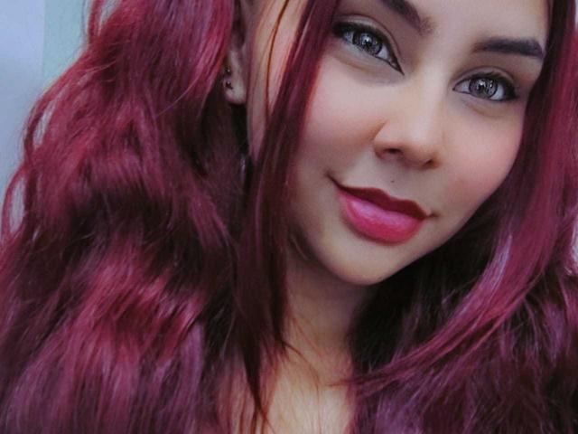 JulietteQueen mit roten, gewellten Haaren, blauen Augen und pinkem Lippenstift, lächelnd in die Kamera, wobei sie ihre niedliche Nase und ihre kleinen, süßen Brüste zeigt, mit einem subtilen, sexy Blick..