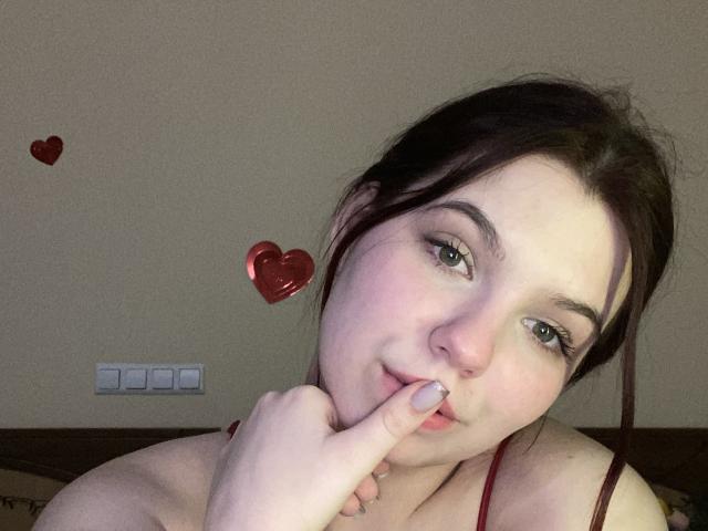 Khaleessi with fair skin, dark brown hair in a messy bun, biting her finger, wearing a red strap, green eyes, heart stickers on the wall, and a light switch.