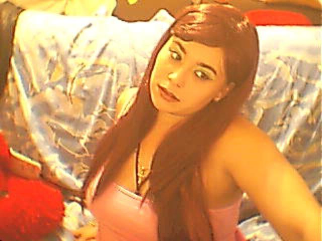 MayaSmith with long brown hair, pink tank top, and a cute expression, holding a bottle, has medium-sized tits, and a soft, warm light on her face.