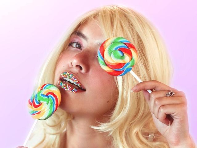 LilibethHot69 con i capelli biondi, lecca-lecca colorati a spirale davanti e dietro al suo viso, labbra arcobaleno scintillanti, che tiene un lecca-lecca, indossa un anello, sfondo rosa sfumato..