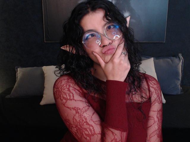 LilibethHot69 con capelli neri ricci, orecchie a punta, e occhiali con montatura blu, che fa la smorfia, con le mani sul viso, indossa una camicetta rossa in pizzo, stanza buia, cuscino e quadro appeso al muro..
