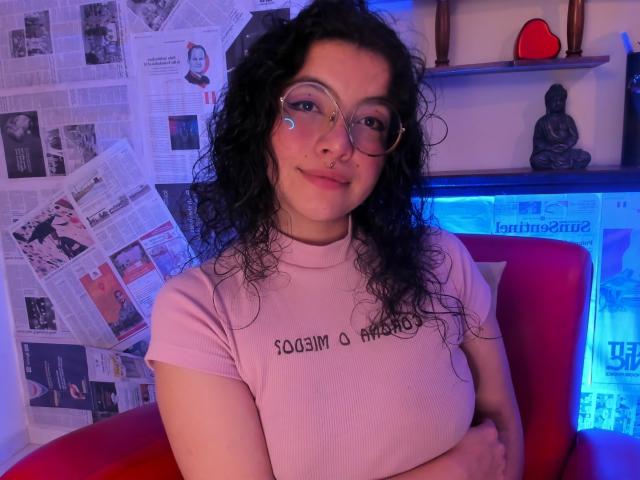 LilibethHot69 mit lockigem, schwarzem Haar, Brille, Nasenring, rosa Hemd, verschränkten Armen, sitzend auf einem roten Stuhl, blaues und violettes Licht, süßes Lächeln, kleiner, blauer Herz-Sticker auf der Wange..