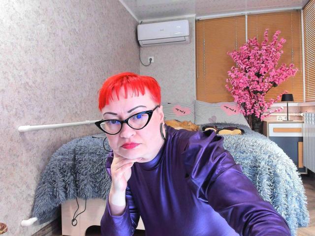 Lackengel-hot with short red hair, black glasses, and purple satin shirt, resting chin on hand, looking at camera. Pink cherry blossom tree in background.