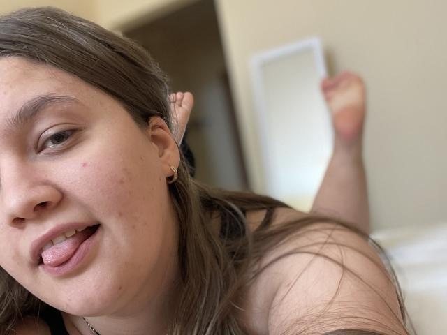 Madrinia's face, brown hair, slightly wavy, with small earrings, sticking out her tongue, showing small zits on her cheek, lying on a bed with her bare ass up.