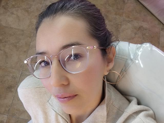 AntonnellaS with clear, round glasses, pink lips, and hoop earrings, wearing a beige sweater, sitting on a white chair, brown tiled floor, and her hair is tied back.