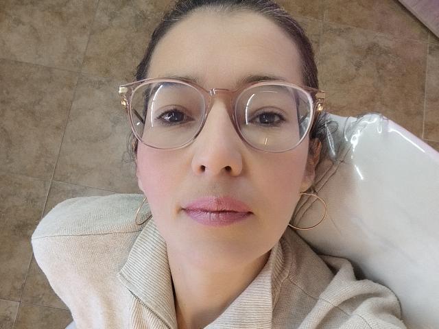 AntonnellaS with glasses, pink lips, and hoop earrings, wearing a beige sweater, looking up with a slight smile, brown tiles in the background.