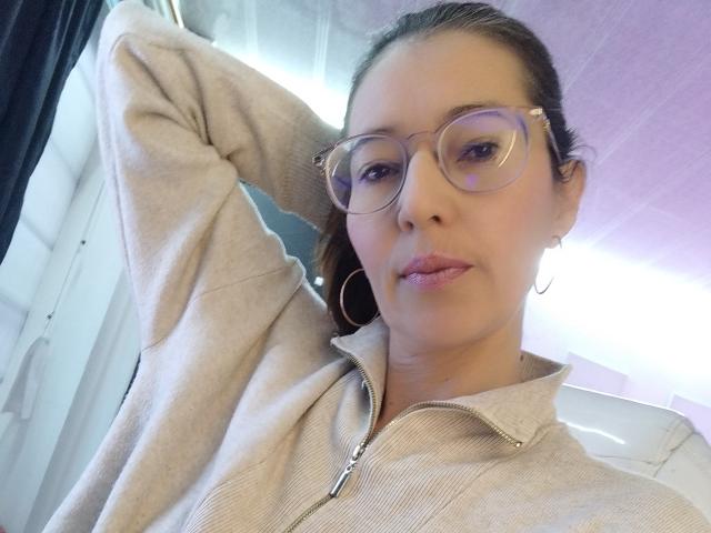 AntonnellaS with glasses, light brown skin, pink lipstick, and hoop earrings, wearing a beige zip-up, hand in hair, looking at camera. Her tits are small, she's cute.