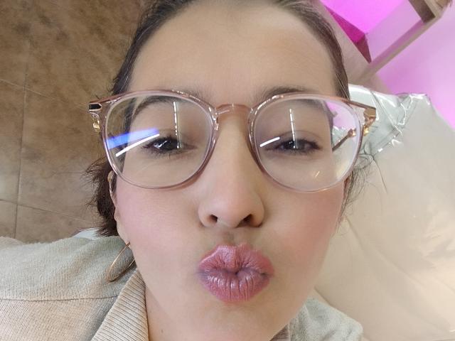 AntonnellaS with pink-tinted glasses, puffy pink lips in a pout, light brown skin, and brown hair, wearing a beige top, reflecting lights in her glasses.