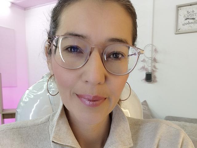 AntonnellaS has shiny pink lipstick, wears large clear glasses, and has hoop earrings, with a beige collared shirt, in a selfie showing her smiling face.