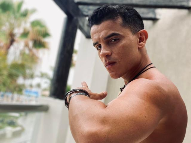 Rocko27 has a muscular, tan, shirtless upper body with defined arms, short black hair, and a black necklace. He's looking over his shoulder with a serious expression.