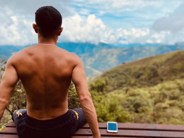 Rocko27, shirtless with a muscular back and brown skin, sits on a wooden bench, facing a mountain view, wearing black pants, with a smartphone on the bench.