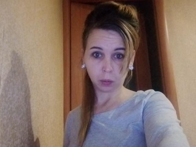 LoveisHt with pouty lips, light brown hair in a half-up style, wearing a gray shirt, small earrings, and a slightly confused expression, standing in a doorway.