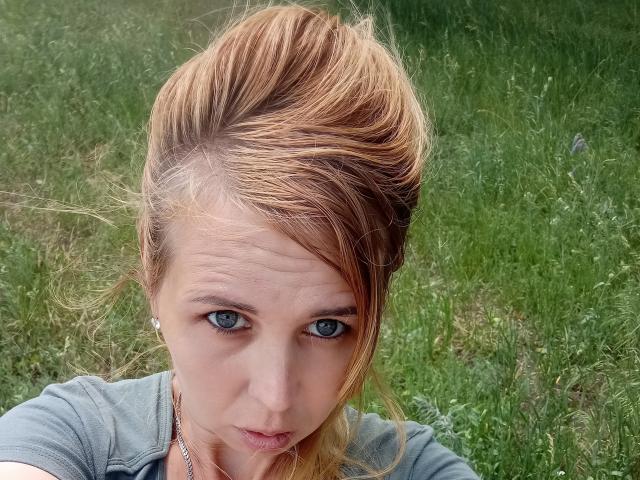 LoveisHt has dirty blonde hair in a messy updo, blue eyes, and a serious look, wearing a gray shirt with a silver necklace, in a grassy outdoor setting.