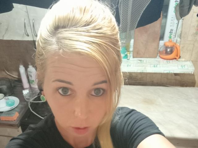 Blond LoveisHt with large eyes, slightly parted lips, and black shirt, holding camera in bathroom, reflecting in mirror. Her ass is barely visible in reflection.