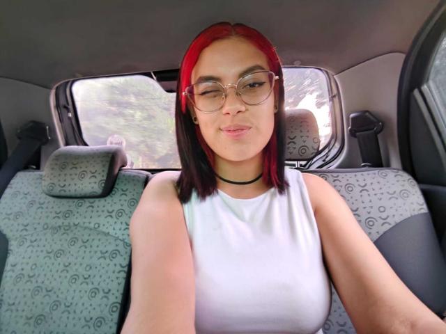 MalejaI with red and black hair, glasses, white sleeveless top, black choker, sitting in a car, smiling, showing small tits, patterned car seats, holding phone for selfie.