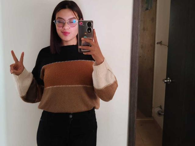 MalejaI has brown hair, glasses, and is wearing a brown and black striped sweater with high-waisted black pants, making a peace sign with her right hand in a mirror selfie.