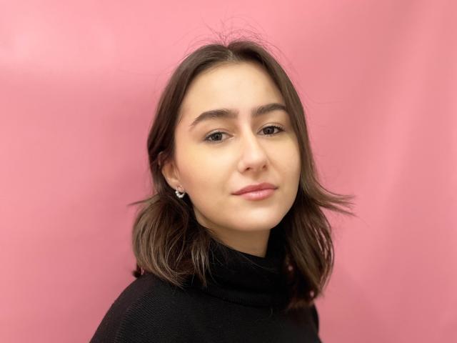 KamilaHarison has shoulder-length brown hair, wearing a black turtleneck, with subtle earrings, and a soft smile on her face, against a pink background.