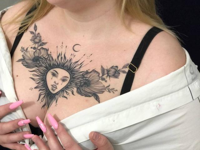 DiLaria's sun tattoo with a face in the center, surrounded by flowers, on her chest, showing black bra straps and pink nails, with a white shirt partially off.