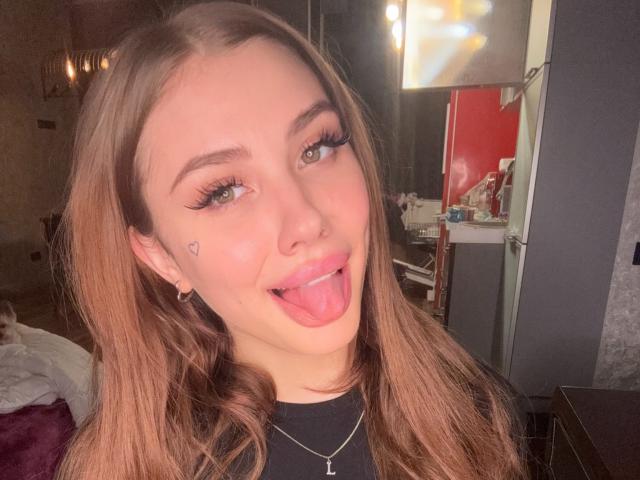 SensualMila a de longs cheveux bruns, des yeux verts, et elle tire la langue en affichant un petit tatouage en forme de cœur sur sa joue, elle porte une chemise noire et un collier..