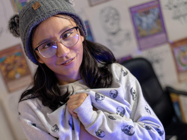 EviieGothel with glasses, gray beanie, pigtails, white skull sweater, holding it, smiling, medium tits, light skin, black hair, office chair in background, colorful posters on wall.