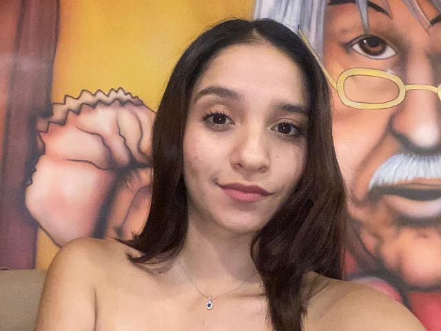 Mabel Prado, com cabelo castanho, pele clara e lábios rosados, ombros nus, com um pequeno mamilo visível, um sorriso discreto, usando um colar fino, em frente a uma pintura colorida na parede..