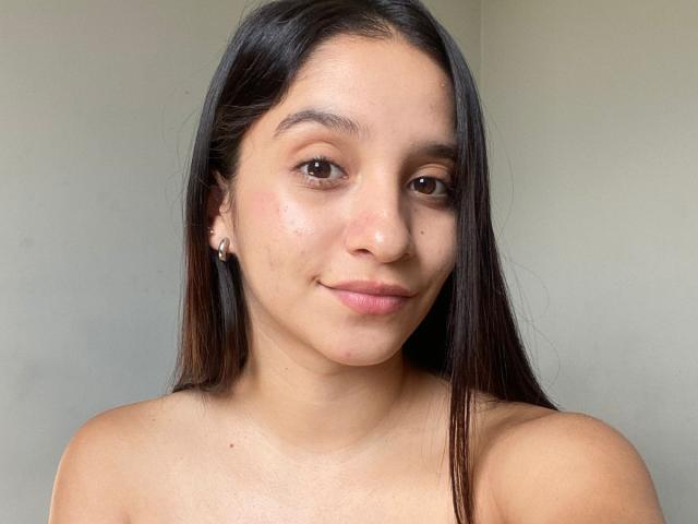 Mabel Prado tem cabelos longos e castanho-escuros, olhos castanhos e um sorriso discreto, com os ombros descobertos, revelando um rosto natural e adorável, com uma pequena pinta na bochecha direita..