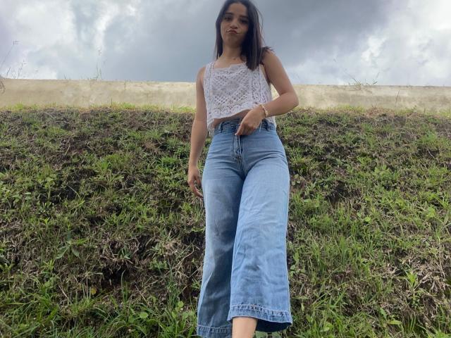 Mabel Prado, con i capelli scuri, una canottiera di pizzo bianco e jeans blu a vita alta, è in piedi sull'erba, guardando in basso, con un cielo nuvoloso sullo sfondo. Le sue natiche sono coperte dai jeans..