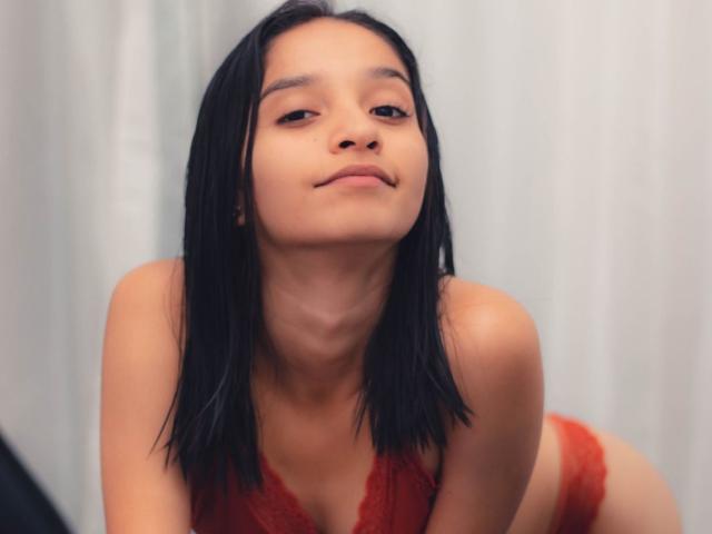 Mabel Prado, avec ses cheveux noirs, sa peau de couleur moyenne et un léger sourire, portant de la lingerie en dentelle rouge, penchée en avant, montrant de petites seins, et une expression confiante et sexy..