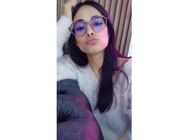 LyraRaven with glasses, black hair, pinkish lips, white fluffy shirt, gray leggings, hand in hair, pinkish-blue reflection in glasses, sitting, relaxed, slightly smiling.