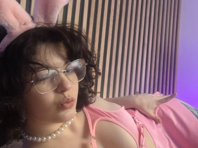 CurlyVixen with curly black hair, pink bunny ears, glasses, pearl necklace, pouting lips, pink dress with cutouts, hand on breast, striped background.