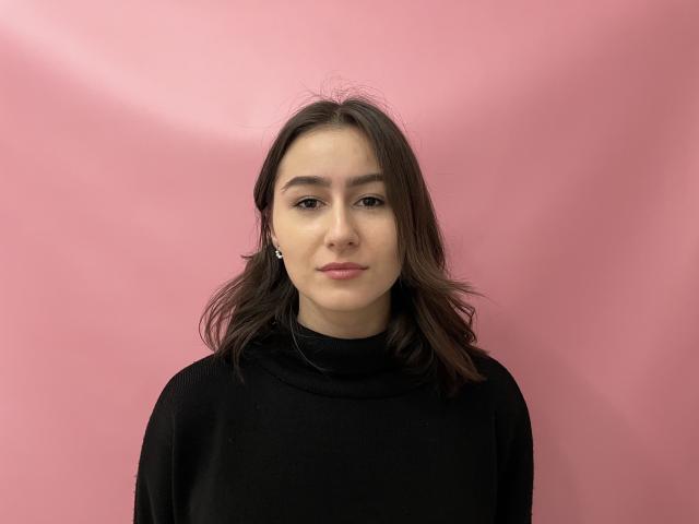 KamilaHarison with medium-length brown hair, wearing a black turtleneck, has a calm expression, and is looking directly at the camera. Her ass is not visible.