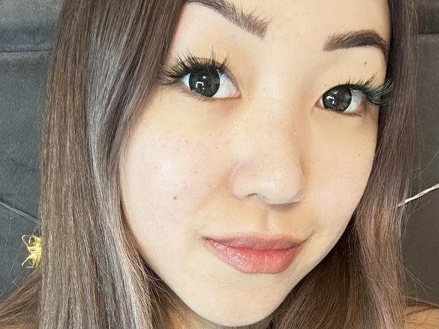 RenyLime's face, showing her green eyes, long brown eyelashes, pink lips, and light skin with some freckles, against a gray background. Her brown hair is straight.
