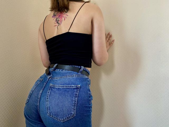 Swoiia with a black top showing her back, butt in high-waisted blue jeans, and a pink floral tattoo on her upper back, standing against a beige wall.