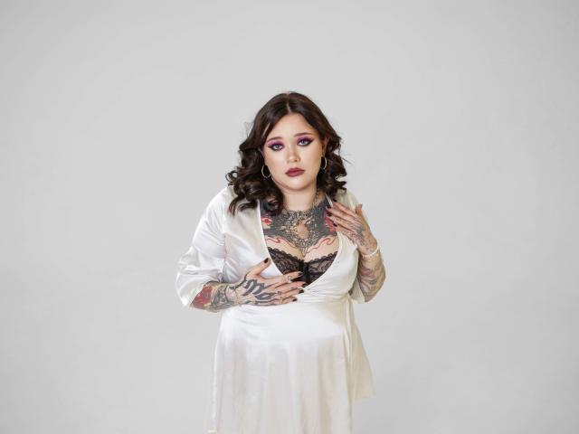 YoureGift with tattoos, wearing a white satin dress, black lace bra, purple eye makeup, dark wavy hair, touching chest, showing tits, holding dress, looking at camera.