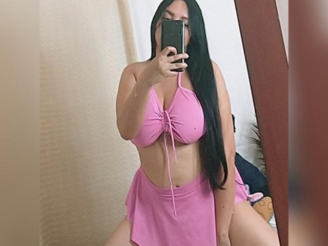 PerlaLovers with long black hair, wearing a pink bikini top and matching skirt, taking a mirror selfie, showing large, prominent tits and a visible, cute ass.