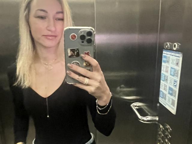 Lilitraya with blonde hair, wearing a black zip-up top, taking a mirror selfie in an elevator, holding a phone with stickers, smiling, showing her tits and ring.