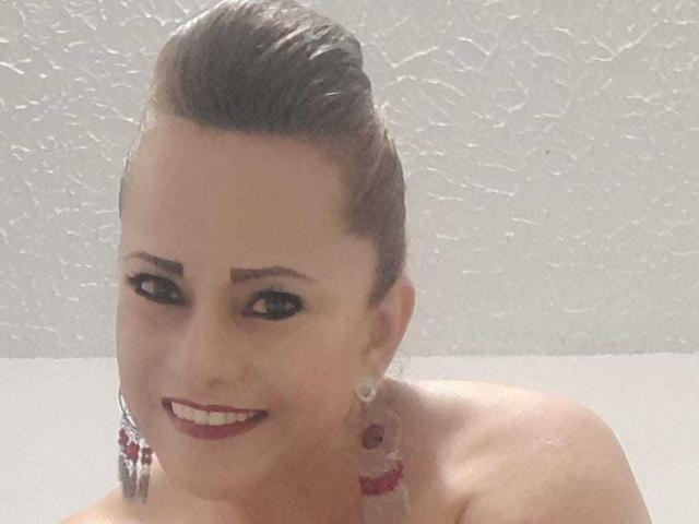 MaryMidori with slicked-back brown hair, wearing red earrings, smiling with red lipstick, showing her bare shoulders and white textured ceiling in the background.