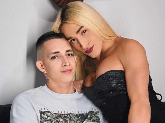 Myluandjachot with short black hair and a white shirt, sitting next to myluandjachot with blonde hair, large boobs, and a black strapless dress, leaning in.
