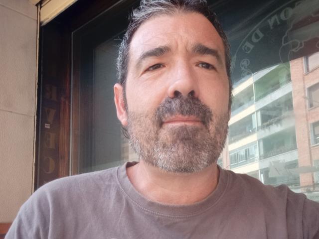 GiovaniFosal has a slightly graying, thick beard, short black hair, and a neutral expression. He's wearing a brown shirt, and his nose is prominently centered.