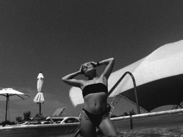 Black-and-white photo of JudyGrace with hands behind head, wearing a dark strapless bikini top and black bottoms, standing by a pool with a large umbrella in background.