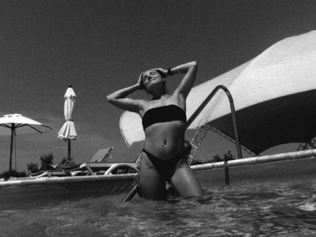 JudyGrace, wearing a black bikini, kneels in a pool with water up to her thighs, hands behind her head, looking up. Black and white, with a large umbrella.