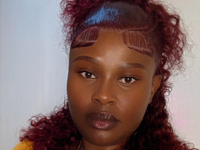 EbonyGoddez has dark brown skin, curly red hair, and straight red bangs, with a serious expression, wearing dark lipstick, and a yellow shirt.