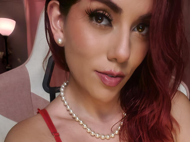 ZoeeMar with red hair, pearl necklace, and red dress, has glossy pink lips, brown eyes, and a slight smile, showing off her pretty face and cute earrings.