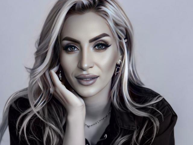 JessycaPearl, con i capelli biondo platino, gli occhi azzurri e le labbra piene, appoggia una mano sul viso, indossa una maglietta nera e una delicata collana, appare sensuale..