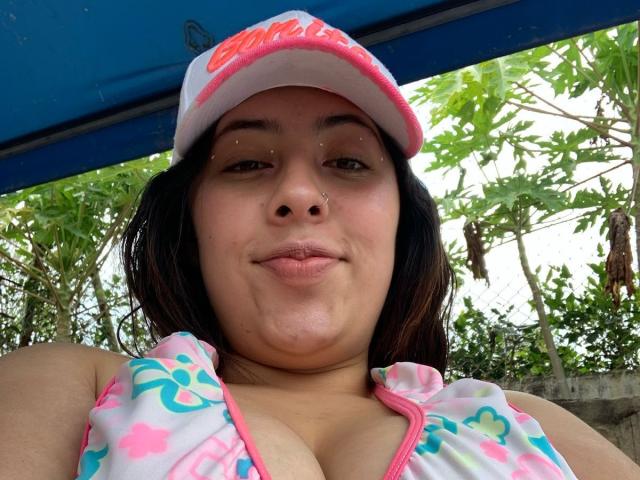 ScarlettParchMusa with medium tits, pink nose, and a cute smile, wearing a white and pink floral top with a matching cap, under a blue canopy, surrounded by green leaves.